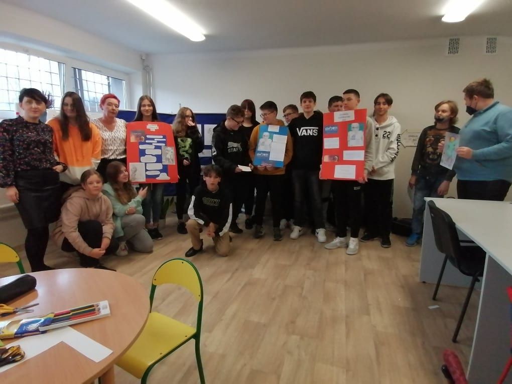 LEM 1- Uczniowie klasy VIII b zgromadzeni w półkolu prezentują swoje plakaty na temat postaci Stanisława Lema wykonane podczas lekcji bibliotecznej.