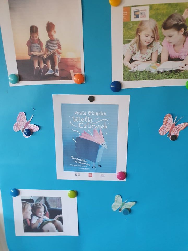 niebieska plansza z przypiętymi zdjęciami dzieci oglądającymi książki. Napis na środku planszy" Mała książka- wielki człowiek"
