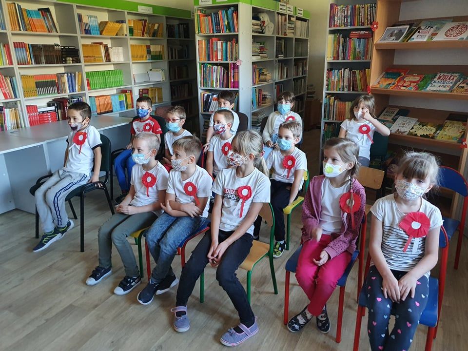 Grupa dzieci z klasy 1b w maseczkach i przypiętych biało czerwonych kotylionach siedzi na krzesłach w bibliotece.